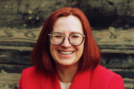 A woman with red hair, grey glasses, and a red jacket smiling