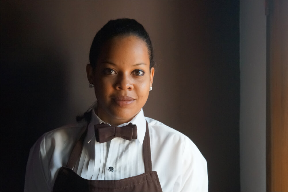 A woman smiling who works in the food industry, with a brown bow tie, white dress shirt, and brown apron