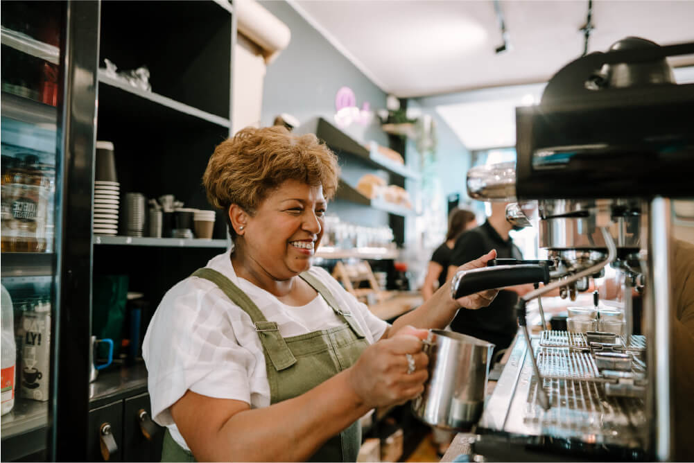 An older woman with short curly hair, smiling, with a green apron holding a metal tin pot to a coffee maker