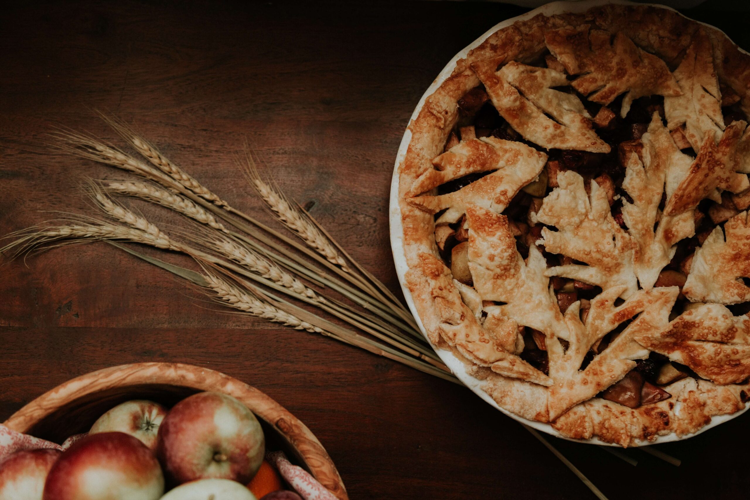 An image of a pie with wheat on the side and apples in a bowl in the bottom left corner