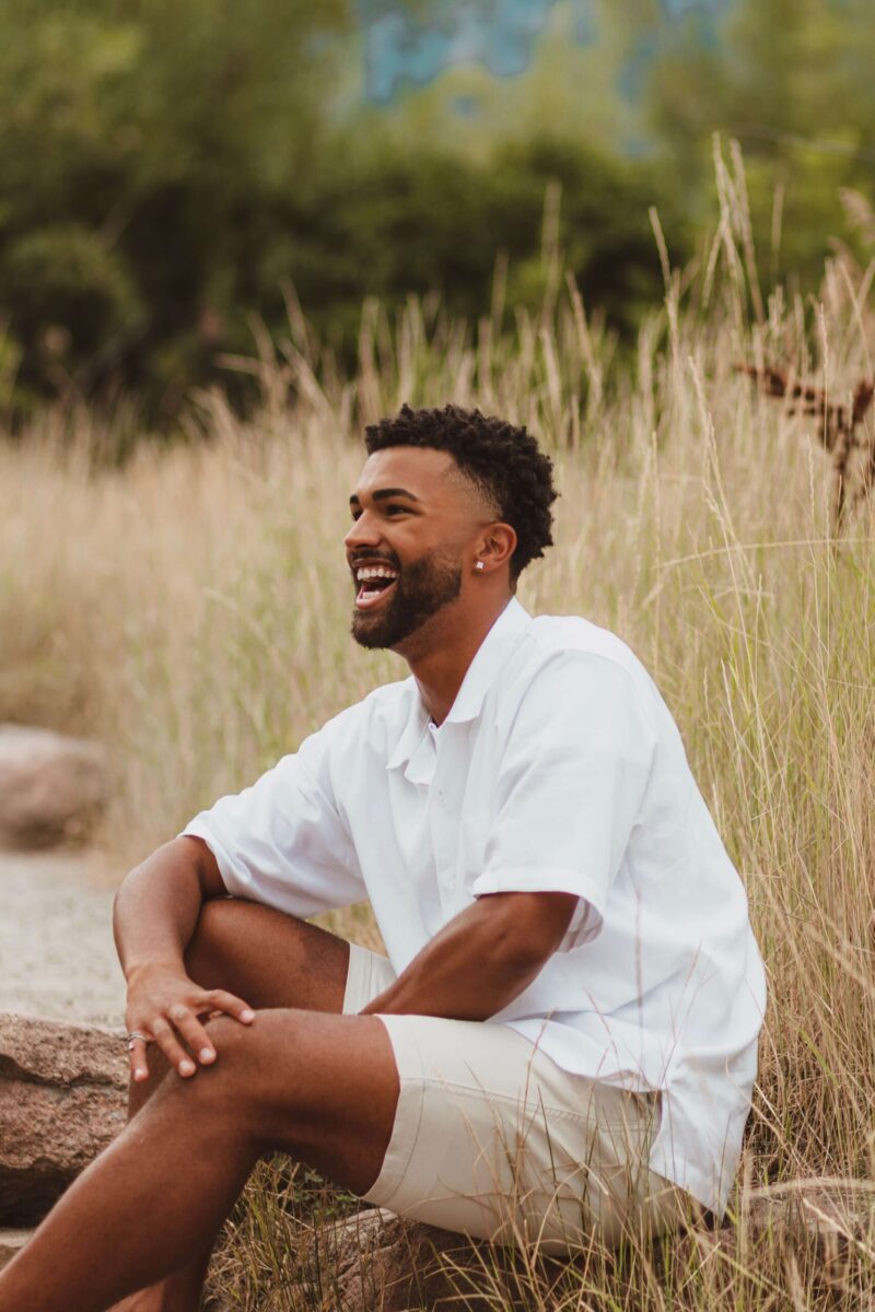 An image of an African American male in white smiling to the left of the frame, sitting on a log